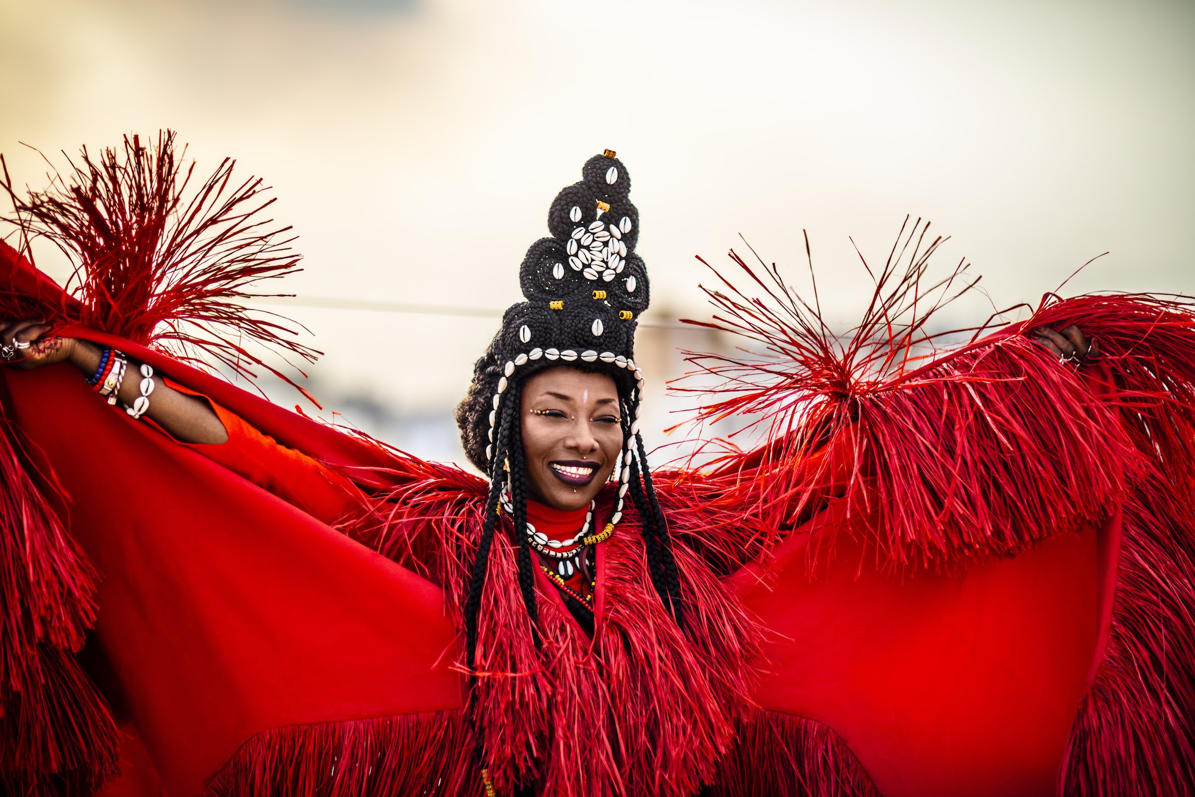 Fatoumata Diawara in a red dress and headpiece made of shells, smiling with her arms outstretched. Photo by Alun Be.