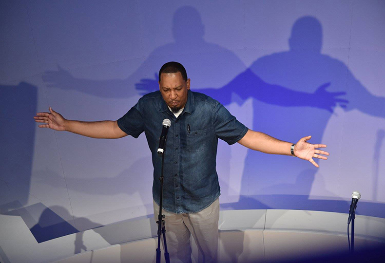 Dasan Ahanu stands with arms wide in front of a microphone.