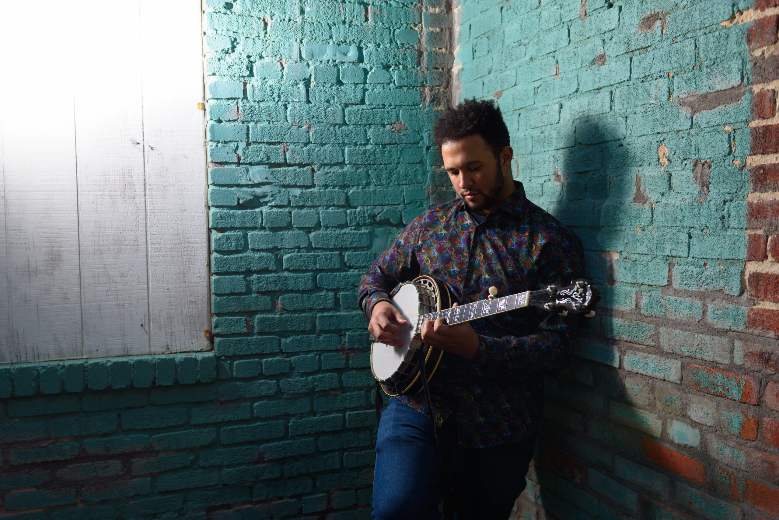 Tray Wellington leans against blue wall while playing a banjo.
