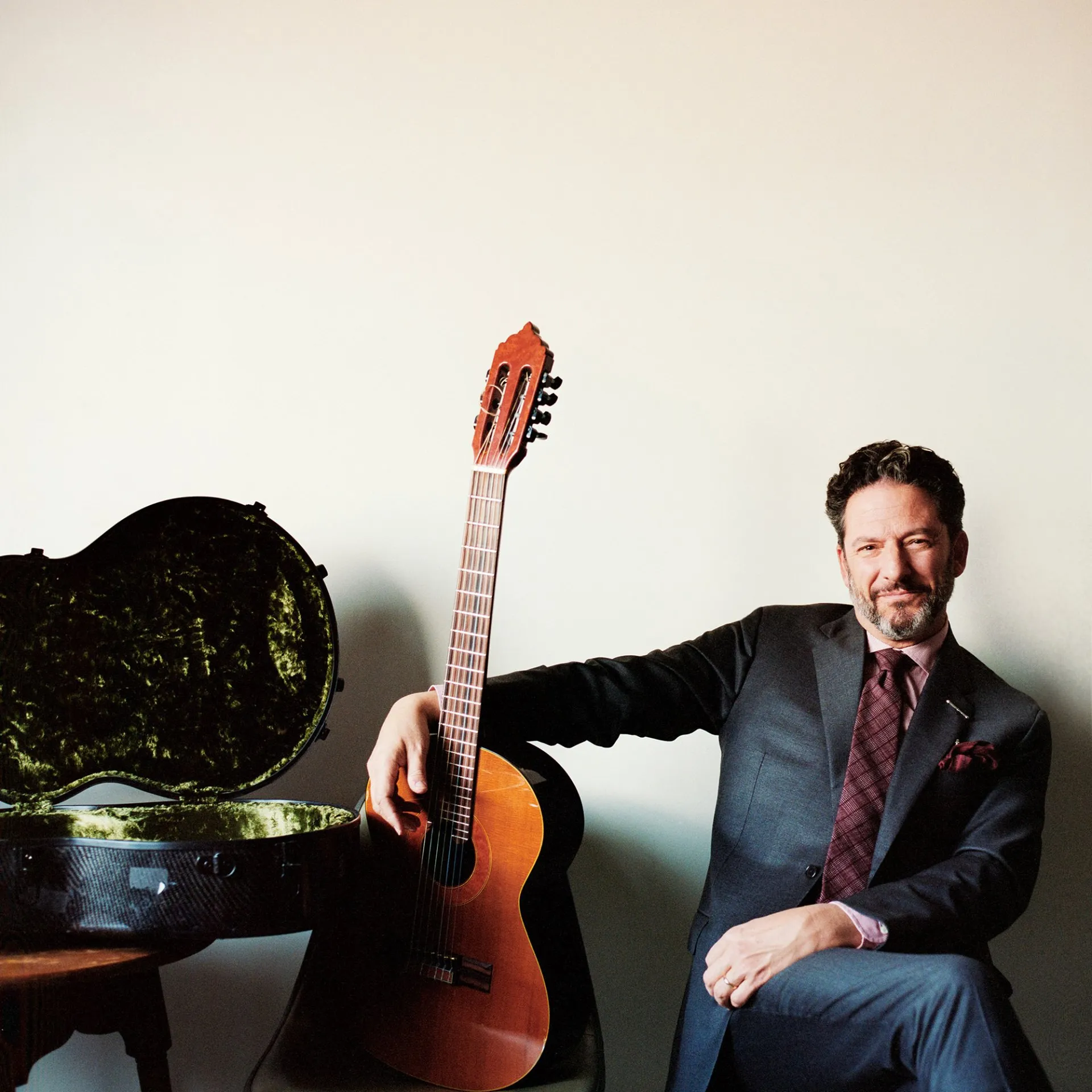 John Pizzarelli sits with his arm around a guitar, next to an open guitar case.