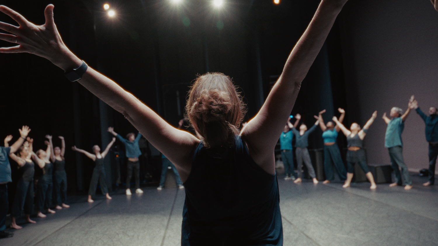 Back view of Michelle Pearson, Founder and Artistic Director of Black Box Dance Theatre, as she stretches her arms wide and leads a circle of performers on stage.